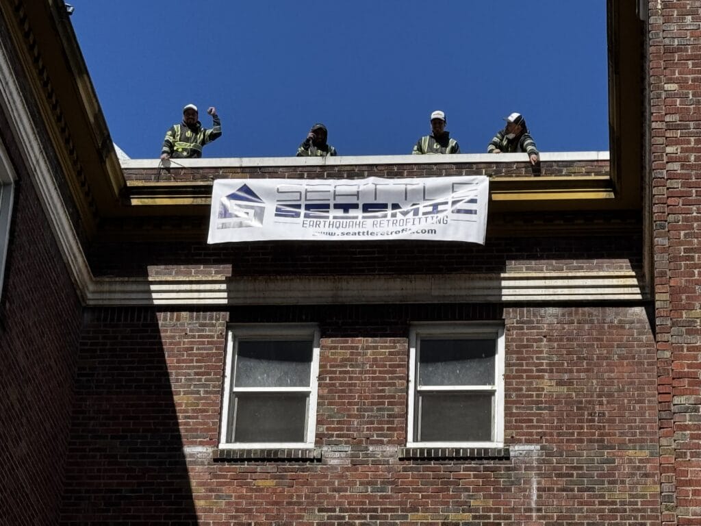 Four people stand on a brick building roof above a white Seismic Earthquake Retrofitting banner, under a bright blue sky.