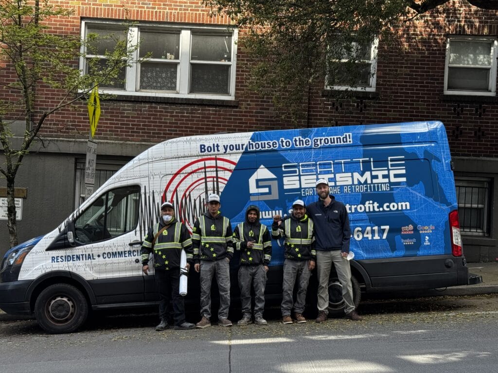 Five workers in safety vests stand in front of a blue Seattle Seismic van parked on a street by a brick building. The van advertises earthquake retrofitting services.