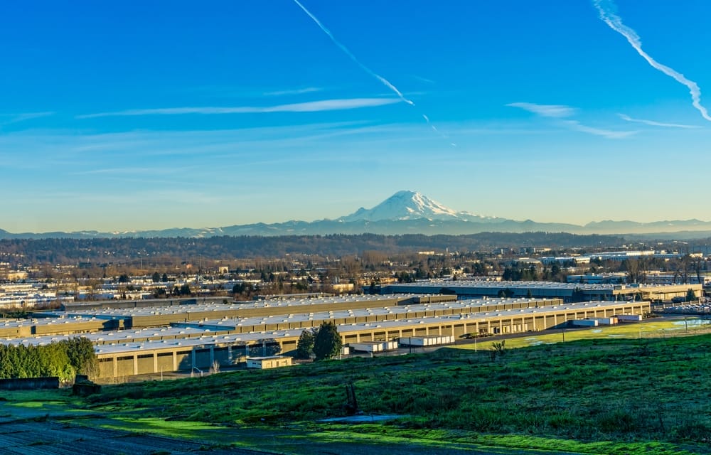 A large warehouse complex sits in the foreground with a grassy field in front and distant mountains under a clear blue sky. A snowy mountain peak rises in the background.