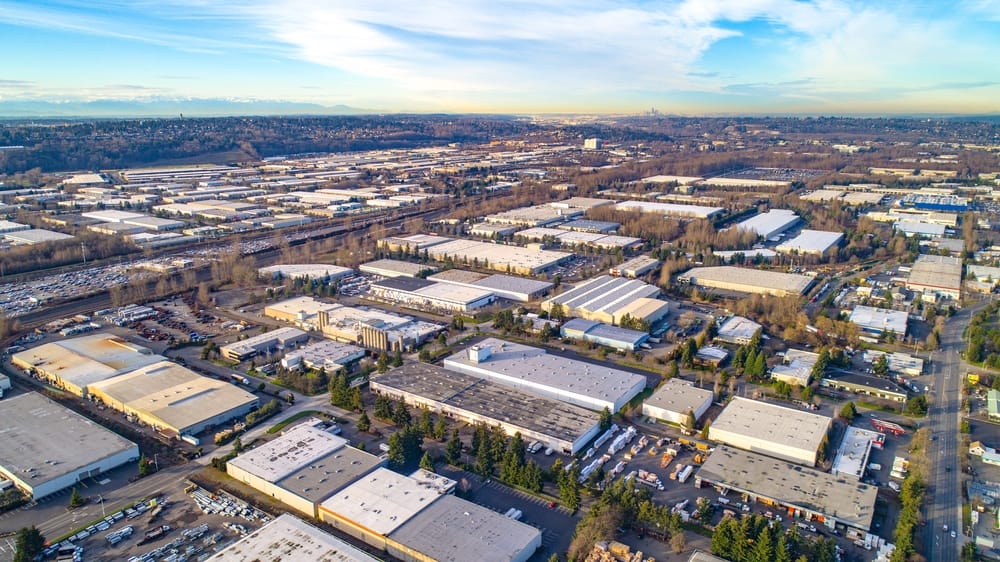 Aerial view of a large industrial area with many warehouses, roads, and some trees, under a partly cloudy sky with distant mountains visible on the horizon.
