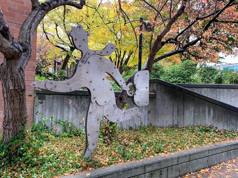 Metal sculpture of a running person holding a briefcase, surrounded by autumn trees with colorful leaves. The sculpture stands on a grassy area near a brick wall and concrete ramp.
