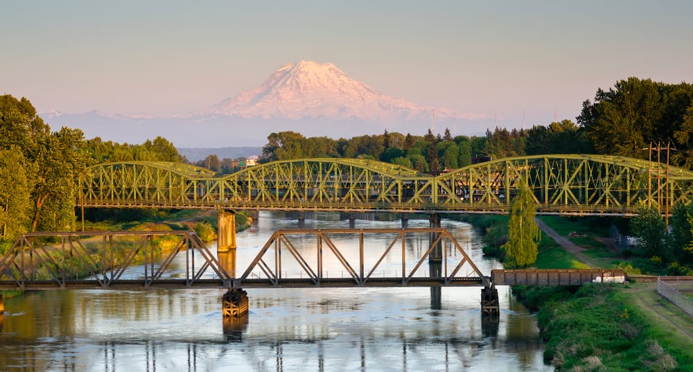 Two green metal bridges span a calm river with lush trees on both sides. Snow-capped Mount Rainier rises in the background under a clear sky.