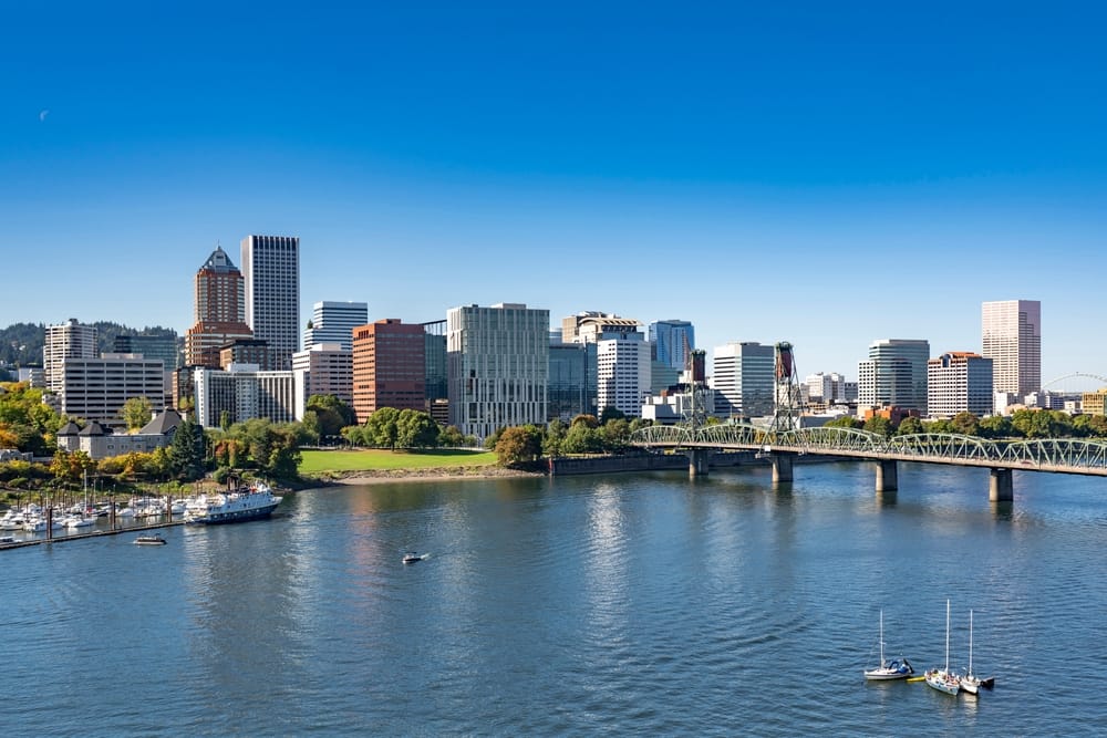 A city skyline with modern buildings, a waterfront, and a bridge crossing a river under a clear blue sky. Boats are docked along the shore, and trees line the waters edge.
