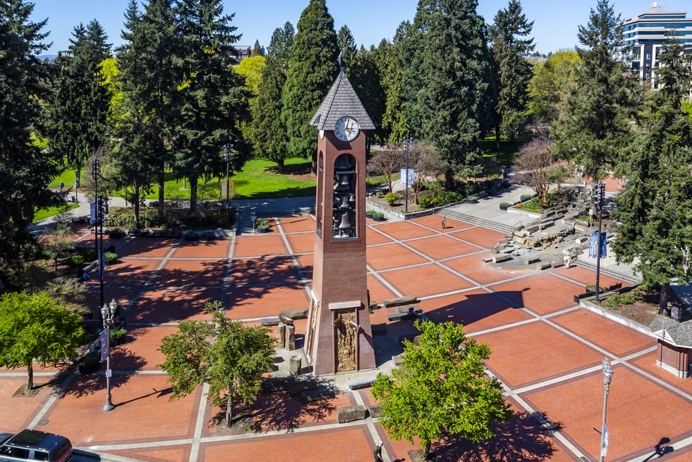 A brick clock tower stands in the center of a large, open square with a red-brick patterned ground, surrounded by green trees and park areas under a clear blue sky.