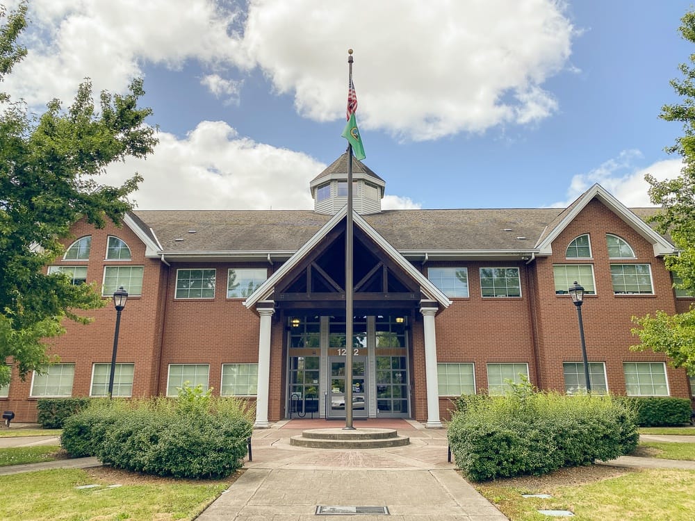 A two-story red brick building with a central entrance, tall windows, and a covered entryway. An American and a green flag fly on a pole. Trees and bushes frame the building under a partly cloudy sky.